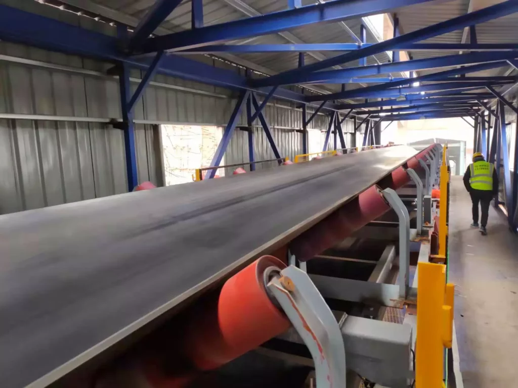 A large industrial belt conveyor with red rollers and a black belt in a steel factory, used for continuous material transport, with a worker inspecting it.